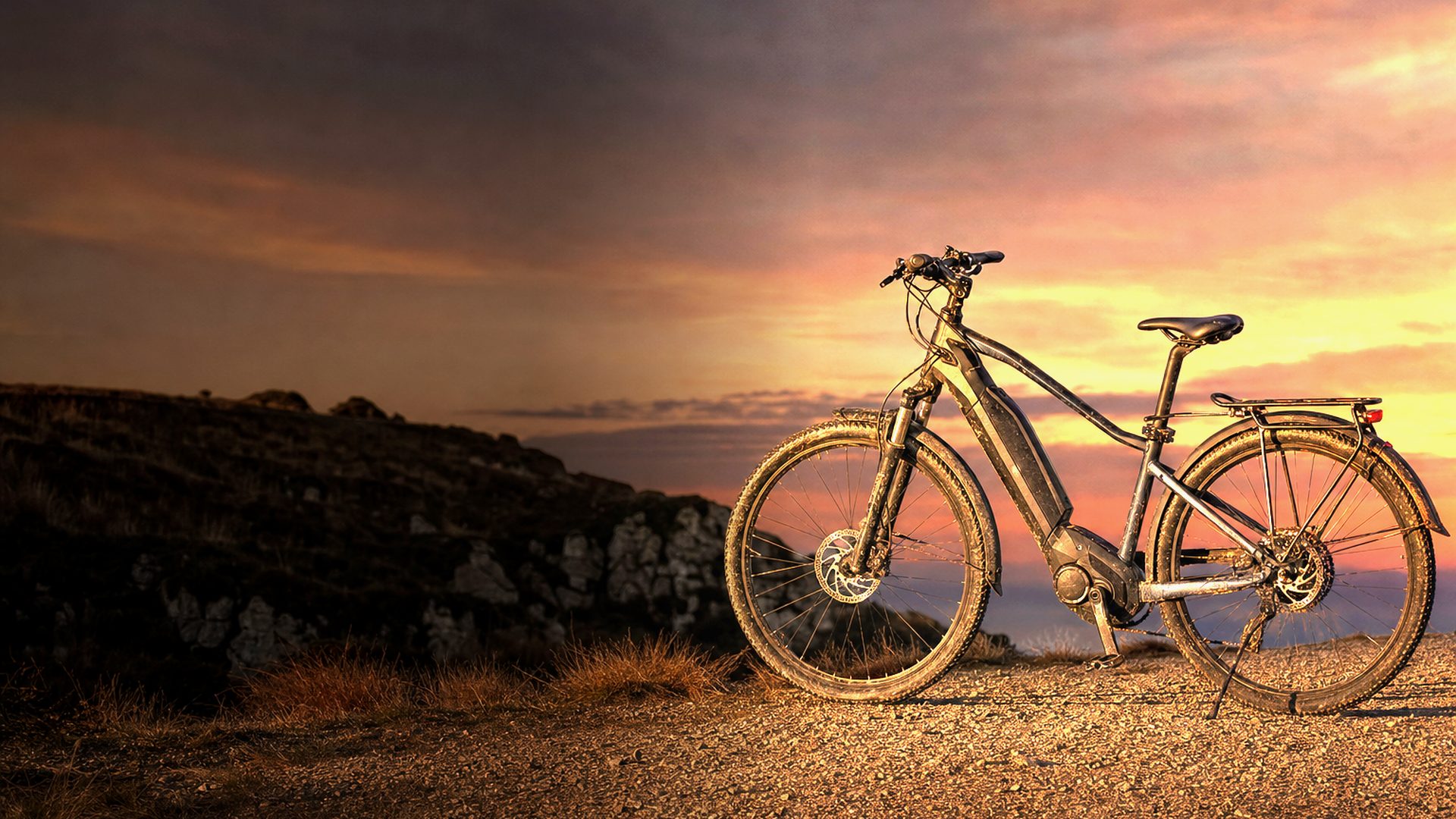 Ein gebrauchtes Trekking-E-Bike steht bei Dämmerung auf einem Schotterrücken, kupferfarbenes Licht bricht hinter dem Rad durch die Wolken
